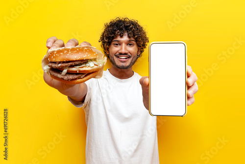 hungry Hindu guy holding tasty big burger and showing blank smartphone screen on yellow isolated background photo