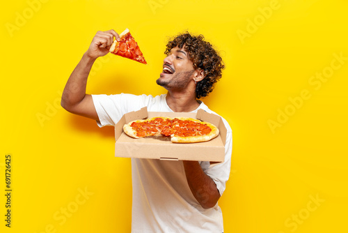 hungry guy indian holds box with delicious pizza and bites a piece on yellow isolated background photo