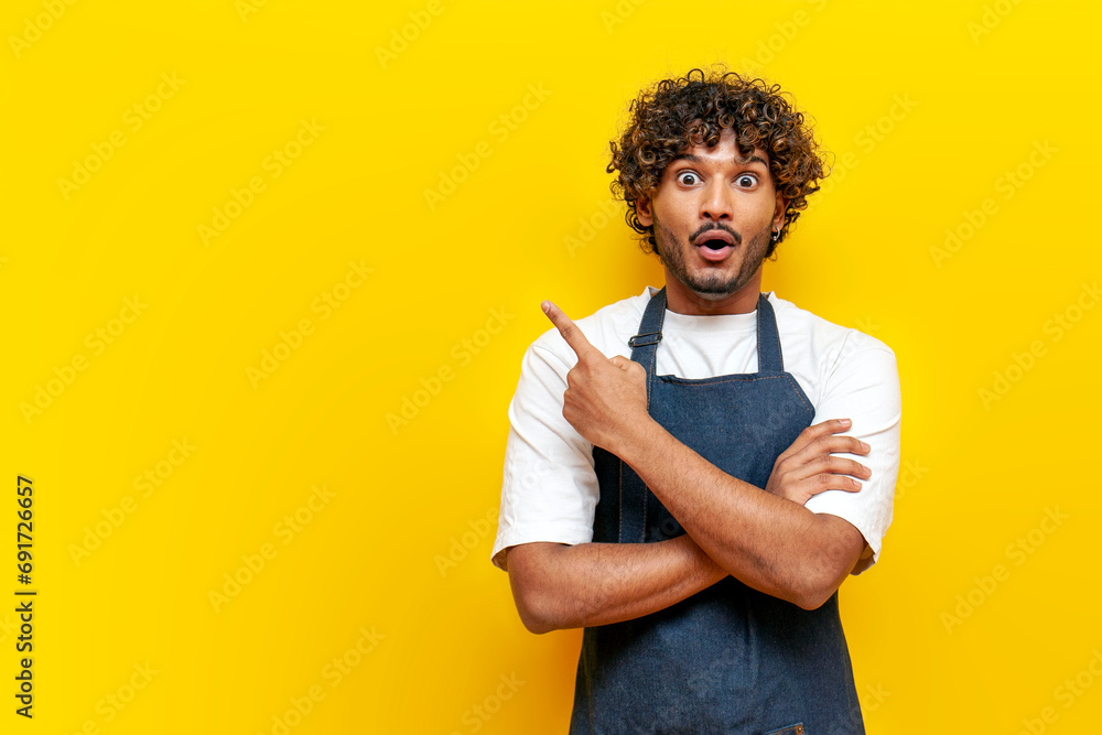 young surprised guy indian waiter in apron pointing at copy space on yellow isolated background - obrazy, fototapety, plakaty 