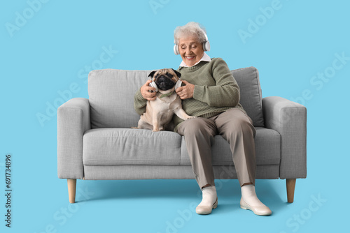 Senior woman in headphones with pug dog sitting on sofa against blue background