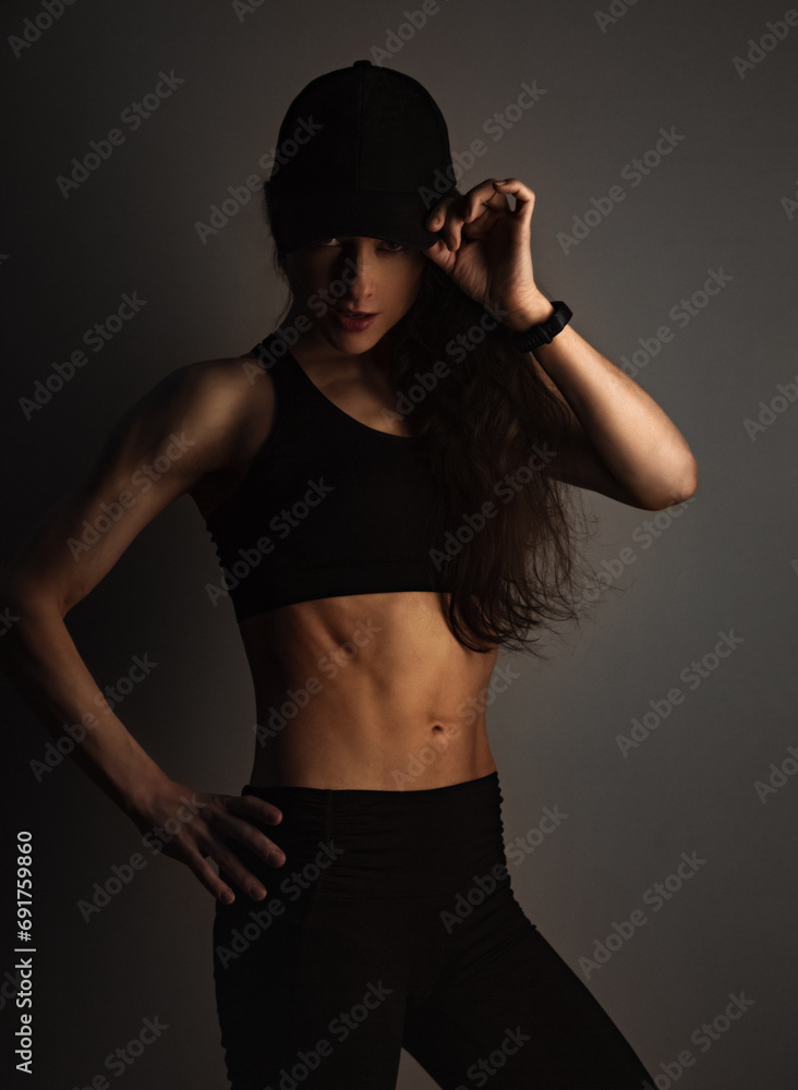 Sport muscular woman posing in black sport bra and cap showing the shoulders, blades and arms, standing on dark shadow studio background. Back body view and profile face. Sporty healthy lifestyle. Sport muscular woman posing in black sport bra and cap showing the shoulders, blades and arms, standing on dark shadow studio background. Back body view and profile face. Sporty healthy lifestyle.
