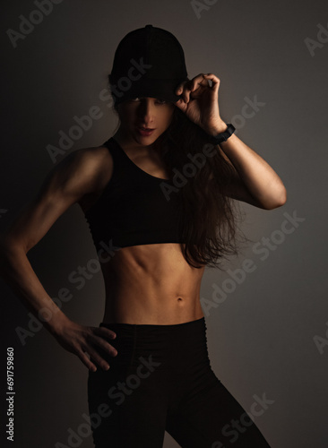 Sport muscular woman posing in black sport bra and cap showing the shoulders, blades and arms, standing on dark shadow studio background. Back body view and profile face. Sporty healthy lifestyle.