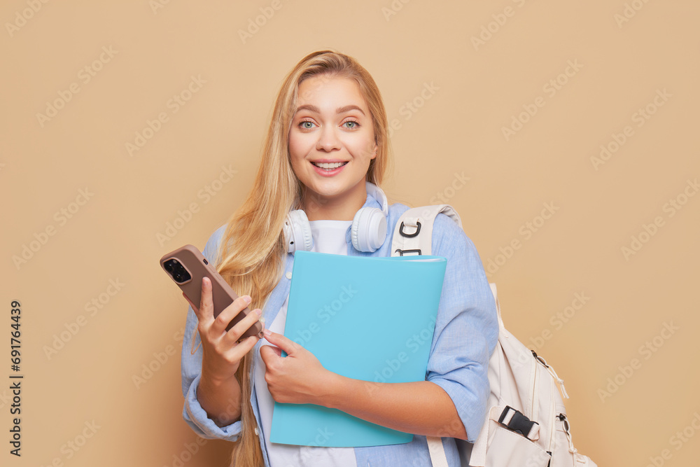 Pretty student girl with long hair holds paper folder and phone in hands smiling happily, college life concept, copy space Pretty student girl with long hair holds paper folder and phone in hands smiling happily, college life concept, copy space