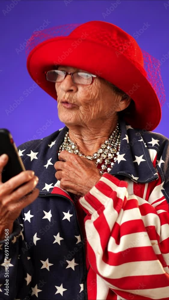 Vertical video of toothless funny old elderly woman with glasses and US flag jacket having video chat on phone smiling and waving isolated on purple background. Concept of crazy senior youthful woman. Vertical video of toothless funny old elderly woman with glasses and US flag jacket having video chat on phone smiling and waving isolated on purple background. Concept of crazy senior youthful woman.