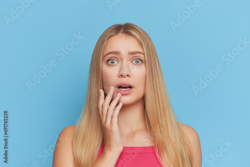 Pretty blonde girl with long hair posing with hand near her mouth over blue studio background, young people concept, copy space