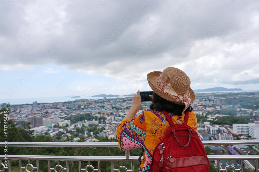 Tourists leaned against the fence and used their mobile phones to take photos of the beauty of the mountains and the misty morning sky. Woman wearing yellow shirt red backpack straw hat and take photo