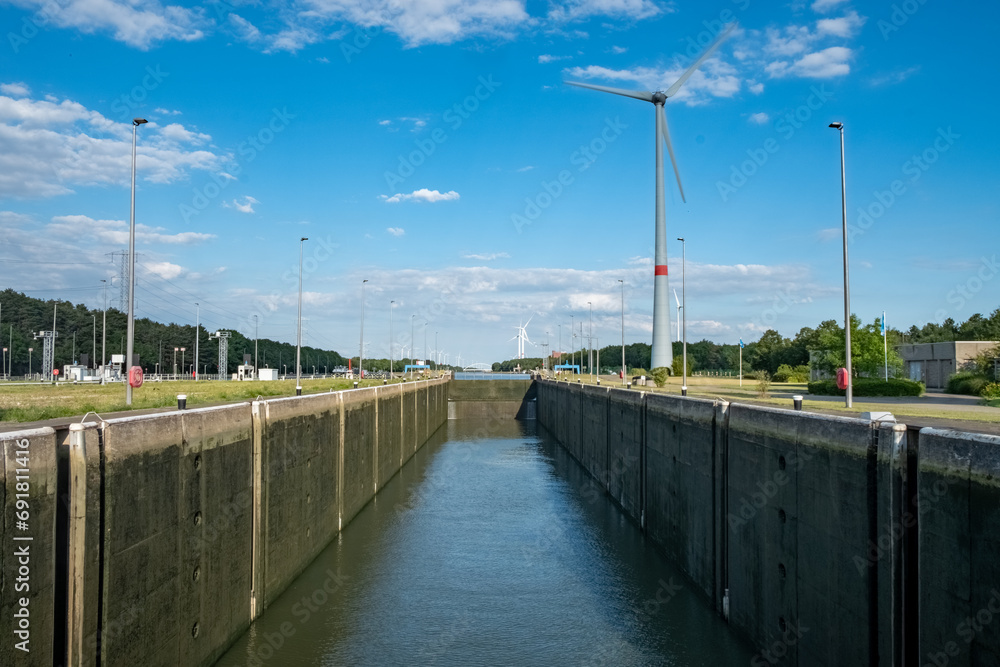 The image displays a spacious lock canal under a clear blue sky, a testament to modern maritime engineering. Flanking the canal, wind turbines stand tall, signifying the harmonious integration of