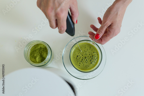 Woman is mixing matcha powder with hot water in glass cup by electric whisk  photo