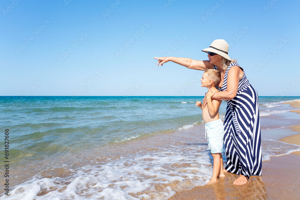 An elderly woman and a boy are standing on the seashore. A grandmother in a long striped dress and hat points her hand into the distance to a grandson in blue shorts. Active recreation.
