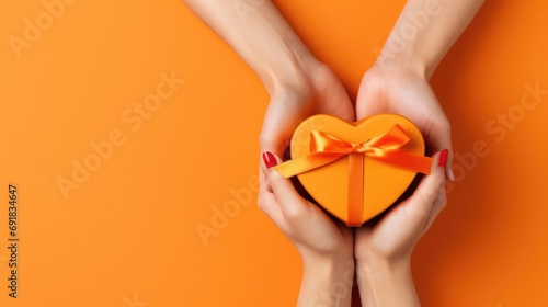 Top view of a woman's hands holding a luxurious gift box with a heart-shaped bow on an orange background.