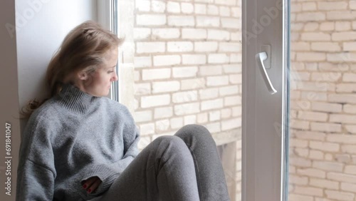 A young tired blonde sits on a windowsill in a cozy warm gray sweater and lets down her hair.