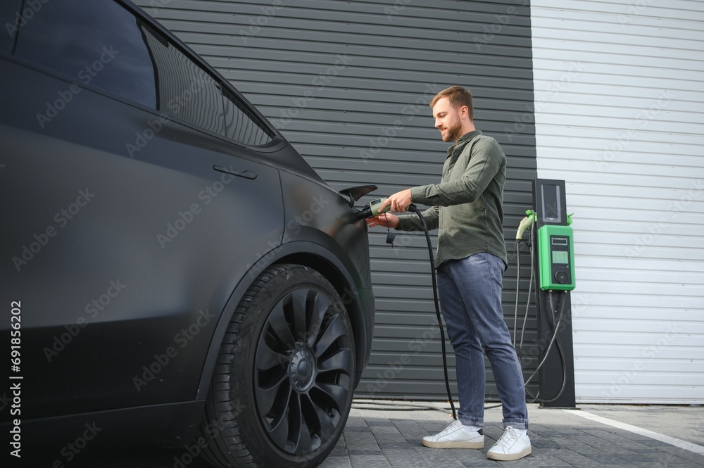 handsome bearded man in casual wear, standing at the charging station and charger for an electric car. Eco electric car concept