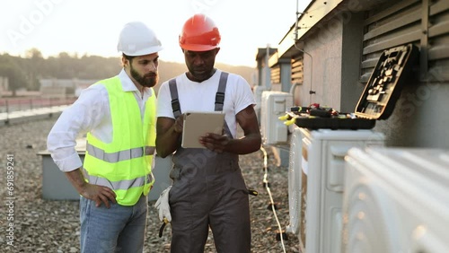 Multicultural males advising with each other about repairing of air conditioner while standing near broken cooling system outdoors. Men using tablet for scrolling websites with useful information. photo