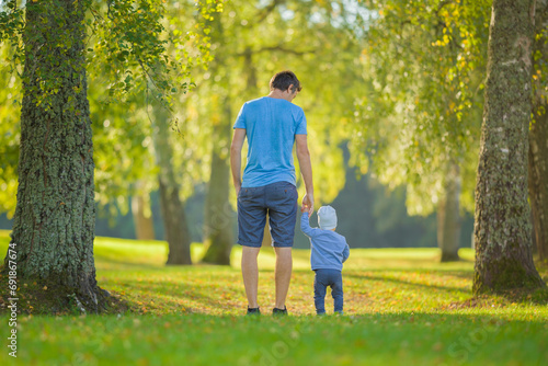 Young adult father and baby boy walking on green grass through tree alley at park. Spending time together in beautiful autumn day. Back view. Lovely emotional moment. Peaceful atmosphere in nature. photo