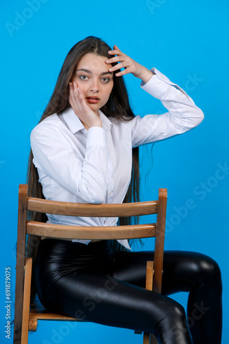 Excited beautiful woman touching face by fingers, while looking at camera. Portrait of worried lady with hands on face, posing, while sitting on chair, isolated on blue background. Concept of posing. photo