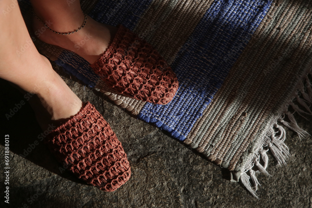 Woman standing in handmade raffia mules on the striped handwoven wool rug. Sustainable ethically made pair of slippers. Responsibly sourced shoes with eco-friendly materials. - obrazy, fototapety, plakaty 
