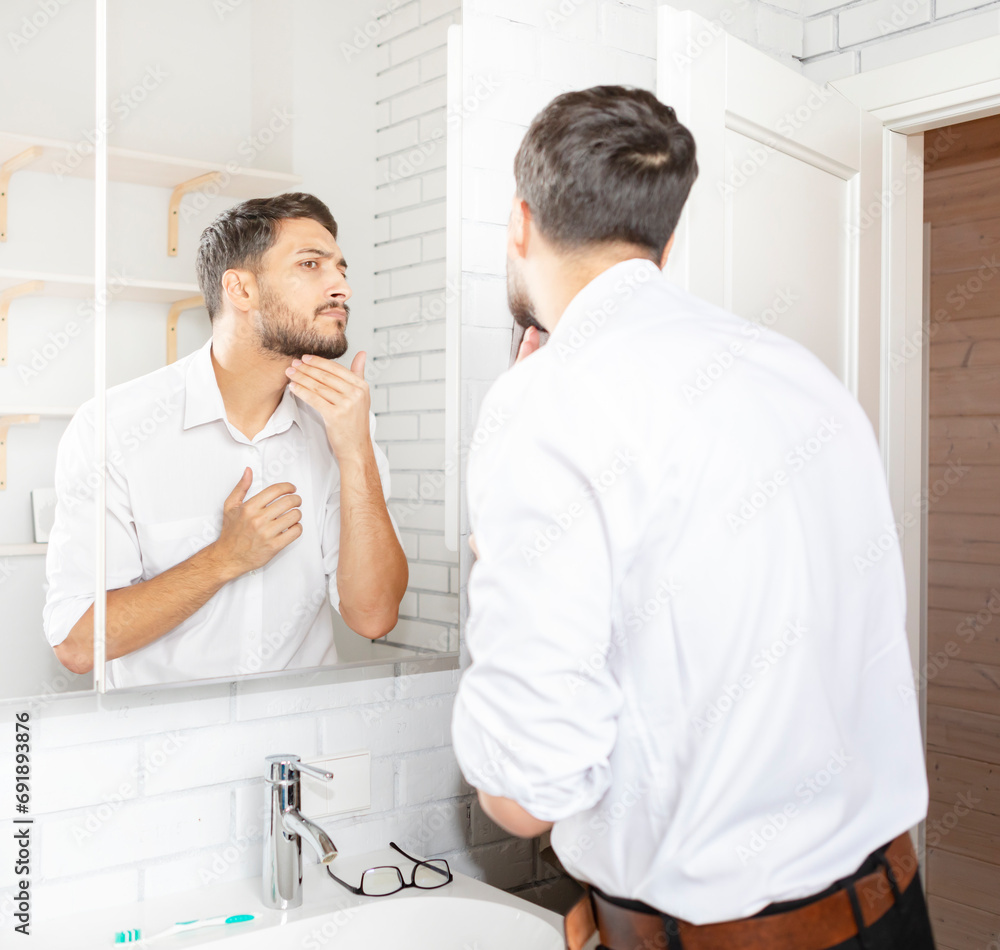 Handsome businessman looking at mirror and applying moisturizing cream on cheeks in bathroom. Groomed young guy doing skincare morning routine after taking a shower Handsome businessman looking at mirror and applying moisturizing cream on cheeks in bathroom. Groomed young guy doing skincare morning routine after taking a shower