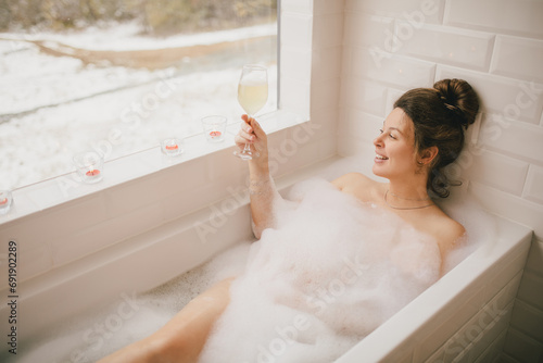 Young woman taking bath with foam near big window.
