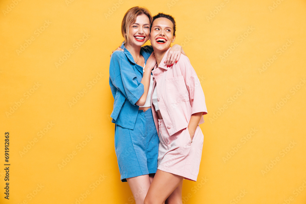 Two young beautiful smiling brunette hipster female in trendy summer clothes. Sexy carefree women posing near yellow wall in studio. Positive models having fun. Cheerful and happy Two young beautiful smiling brunette hipster female in trendy summer clothes. Sexy carefree women posing near yellow wall in studio. Positive models having fun. Cheerful and happy