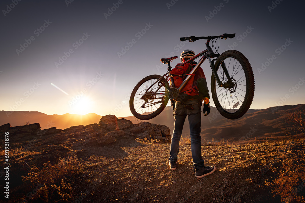 Obraz premium Male mountain biker goes uphill and carry a bicycle on his back to sunset mountain