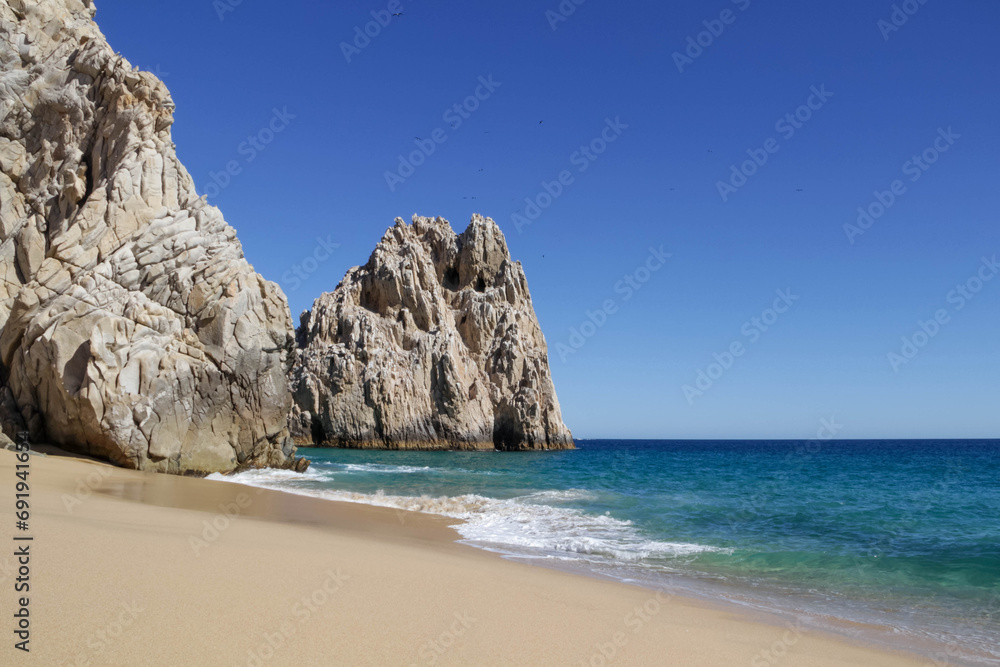 The bright rocks are located on the clear sandy shore of the ocean with water turquoise color. Pacific Ocean; Cabo San Lucas, Mexico.