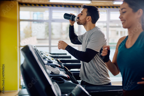 Athletic man drinks water while working out on treadmill in gym.