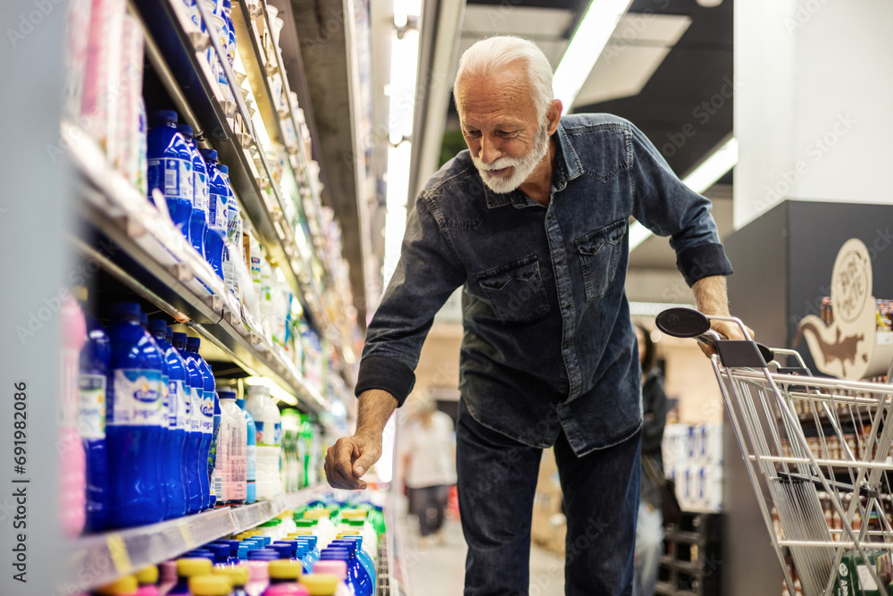 Mature smiling man shopping in local supermarket. Hi is shopping groceries. Walking by refrigerator with dairy product and pushing shopping trolley.