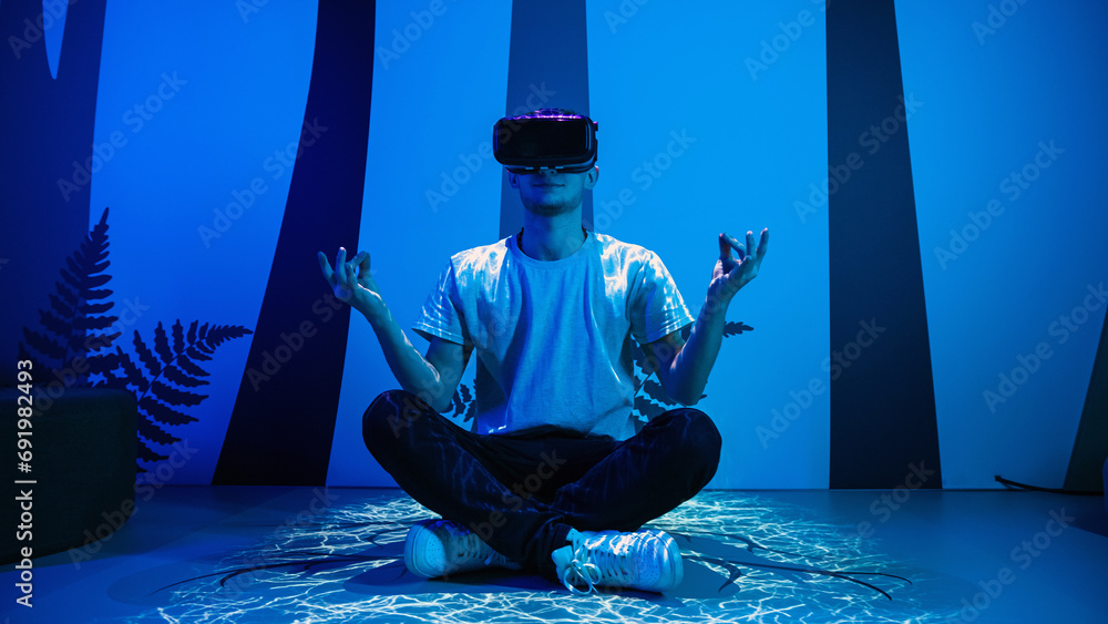 Young man wearing VR glasses sitting in a yoga pose on the virtual reality escape room floor. Fun, entertainment, and innovation concepts. - obrazy, fototapety, plakaty 