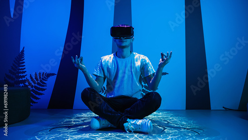 Young man wearing VR glasses sitting in a yoga pose on the virtual reality escape room floor. Fun, entertainment, and innovation concepts. photo