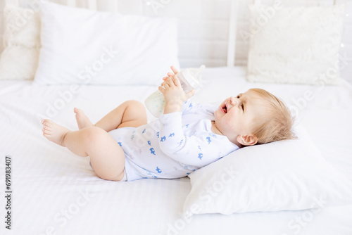 a baby with a bottle of milk does not want to eat and cries holding it in his hands lying on his back on the bed in the children's bright room, the concept of baby food