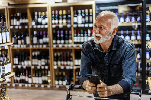 Mature male Caucasian consumer checking information on mobile phone in grocery. Man using cellphone while shopping bottle of wine in the grocery store.