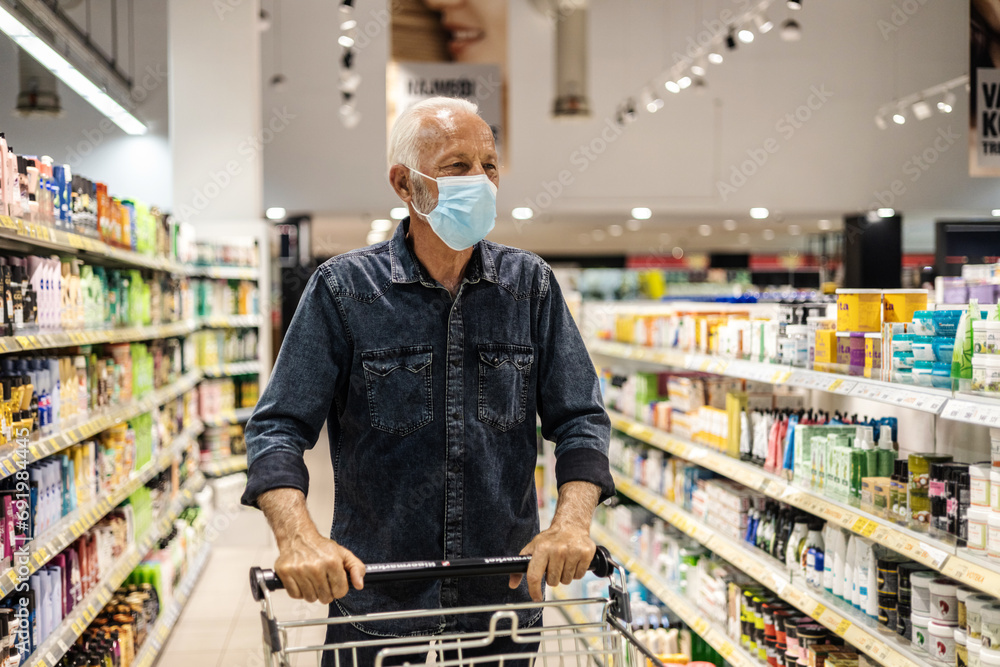 Supermarket, shopping and customer with covid face mask in retail store for food, groceries or product from shelf. Senior man with basket during inflation price increase or sales choice on grocery.