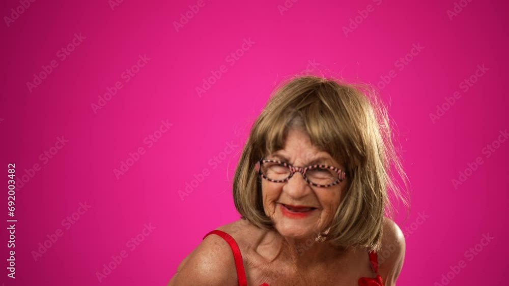Funny elderly woman walks into frame and gives unhappy, angry look at camera. Portrait of old woman isolated on pink background Funny elderly woman walks into frame and gives unhappy, angry look at camera. Portrait of old woman isolated on pink background