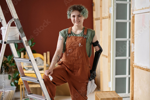 Portrait of young painter smiling at camera while using ladder to paint wall in the room photo