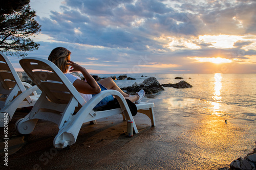 A woman is sitting on a deckchair on the seashore, having fun with her phone and enjoying the sunset photo
