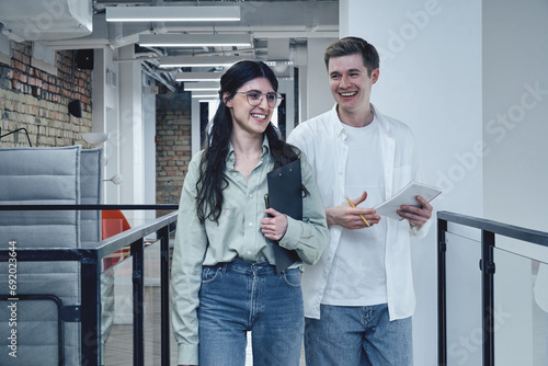 Happy man holding notebook, walking with female colleague with paper holder in the modern office. Teamwork, startup, business concept