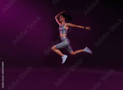 Side view of a slim sportswoman in silver fitness wear jumping against magenta background in studio