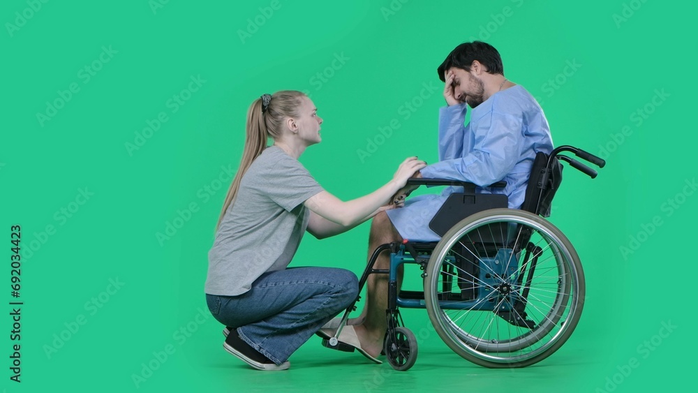 Adult man patient in robe sitting in wheelchair girl next to him holding his hand, supporting expression, man depressed. Isolated on chroma key green screen. Adult man patient in robe sitting in wheelchair girl next to him holding his hand, supporting expression, man depressed. Isolated on chroma key green screen.