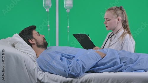 Adult man patient laying in bed with drip, female nurse sitting next to man with clipboard asking questions talking. Isolated on chroma key green screen.