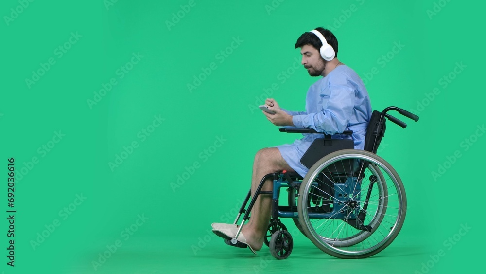 Obraz premium Adult man patient in robe sitting in wheelchair wearing headphones holding smartphone, listening to music. Isolated on chroma key green screen.