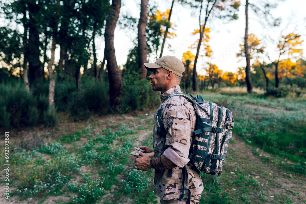 Side view of Army commando with cap and backpack holding box standing in forest Side view of Army commando with cap and backpack holding box standing in forest