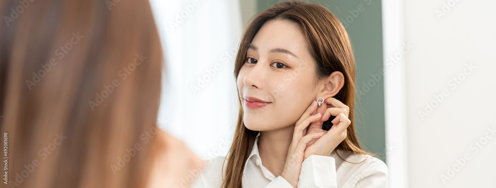 Get dress, asian young woman, businesswoman standing, hand putting earring or jewelry, wearing white shirt formal getting dressed getting ready before go to work looking reflection the mirror at home. Get dress, asian young woman, businesswoman standing, hand putting earring or jewelry, wearing white shirt formal getting dressed getting ready before go to work looking reflection the mirror at home.