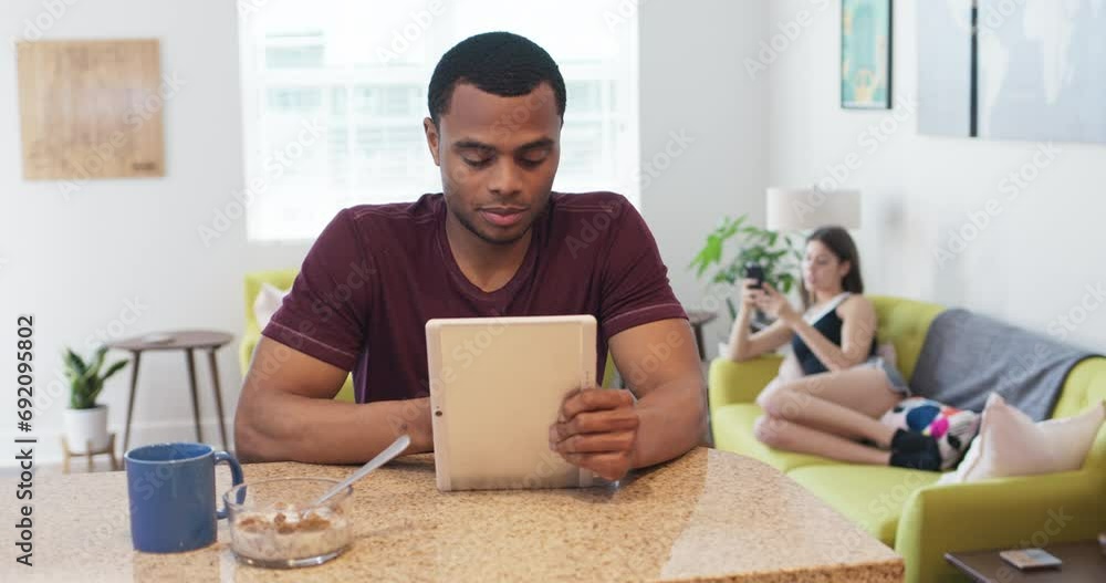 Black millennial man eating breakfast at the kitchen counter while checking his tablet computer with his girlfriend on the couch using her smartphone in the background. 4k slow motion handheld Black millennial man eating breakfast at the kitchen counter while checking his tablet computer with his girlfriend on the couch using her smartphone in the background. 4k slow motion handheld