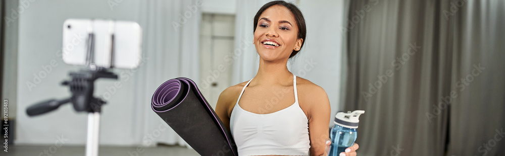 happy african american dancer with fitness mat and sports bottle near smartphone, video blog, banner happy african american dancer with fitness mat and sports bottle near smartphone, video blog, banner
