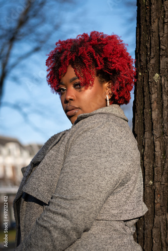 Female model with strong unique style appearance serious moody expression in woodland environment leaning against tree. Confident black lady stylish red hair with winter good taste fashion © drew