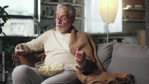 Tired gray haired senior man with remote controller switches channels on TV trying to choose something interesting sitting on couch with bowl of popcorn indoors Boring TV shows films movie  photo