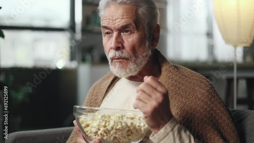Close up portrait of addicted focused senior grey haired man watching interesting TV program film movie and eating popcorn at home Enchanted mature male cannot take eyes off the screen indoors