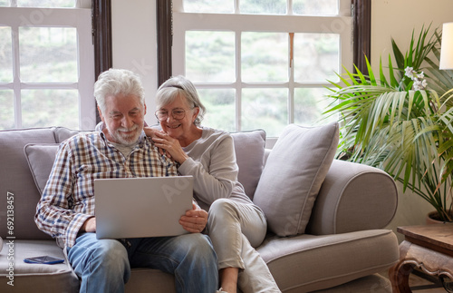 Happy 70s white haired senior couple sitting on sofa in living room using laptop surfing the net. Concept of older generation users and wireless technology © luciano
