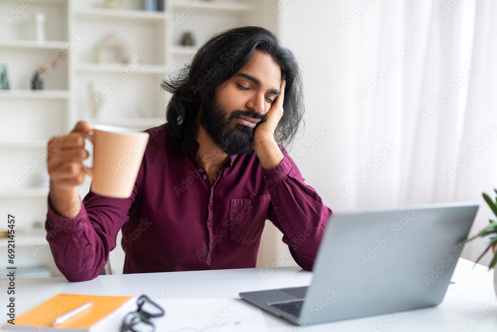 Exhausted Indian Man With Cup Of Coffee Sitting At Desk With Laptop Exhausted Indian Man With Cup Of Coffee Sitting At Desk With Laptop