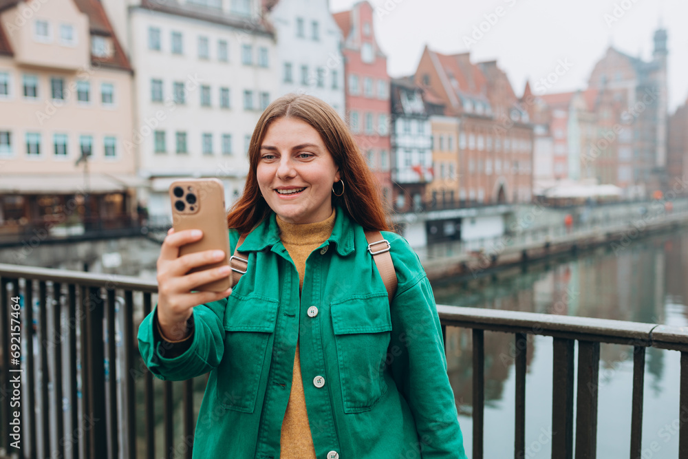 Happy 30s Women taking selfie on urban background. Young beautiful girl say Hi. Gdansk old town and famous Zuraw crane, A beautiful redhead woman is standing near Motlawa river, Traveler